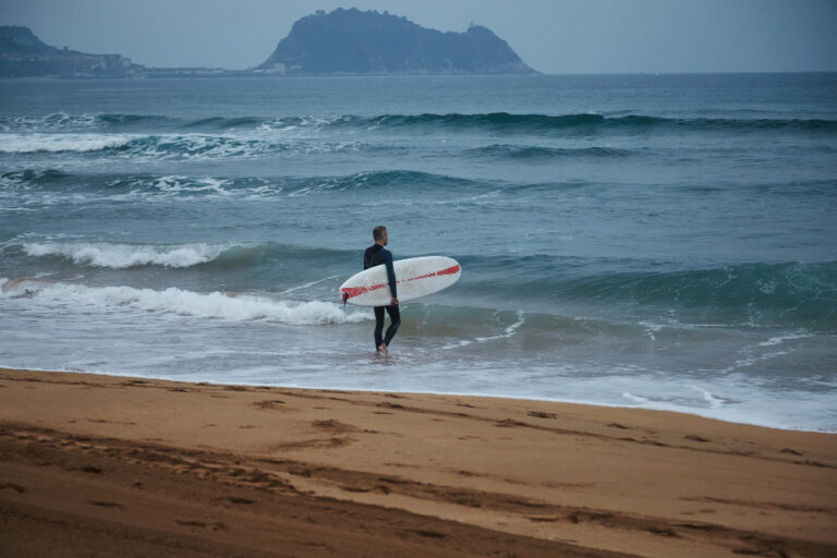 middle-aged-surfer-wetsuit-walking-into-water-sandy-beach-among-hills